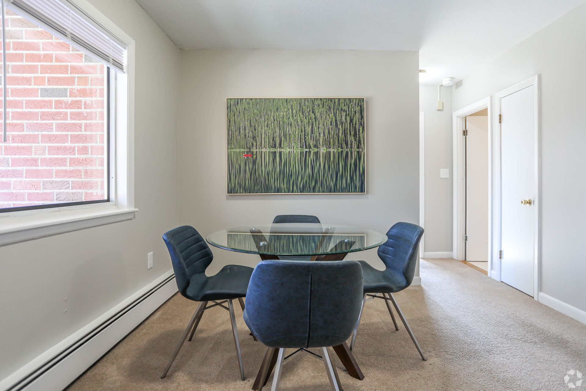 Dining room with glass table, four blue chairs, artwork, and window with brick wall.