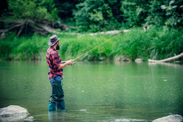 Angler in a red plaid shirt fishing in a calm green river surrounded by lush trees