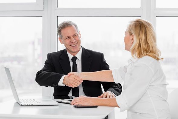 Two professionals shaking hands at a bright office desk with a laptop and documents.
