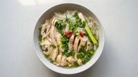 Bowl of noodle soup with sliced chicken, herbs, red chilies, and lime wedges on a light table