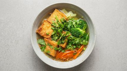 Bowl of noodle soup with tofu, broccoli, and carrots on a gray table