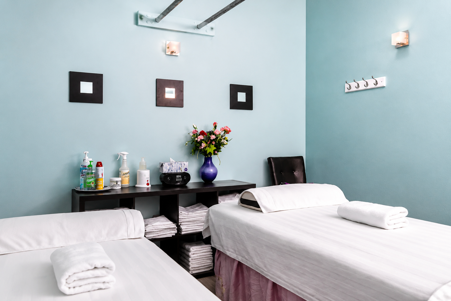 Two massage tables in a spa room with pale blue walls, wall art, and a flower arrangement on a shelf.