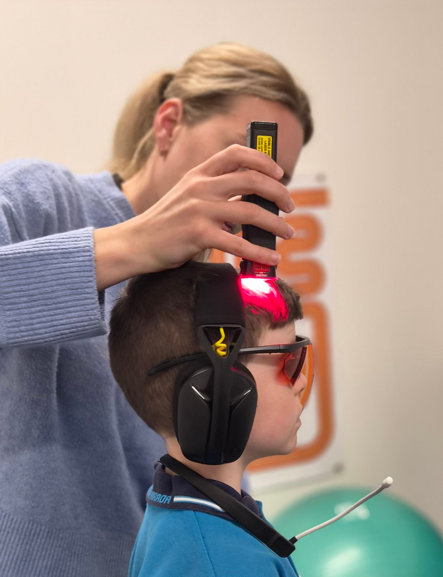 Child in blue sweater wearing a headband while a therapist measures alignment in a clinic with exercise ball background