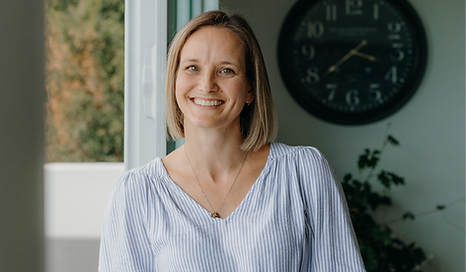 Smiling woman in a striped blouse standing indoors near a window and wall clock