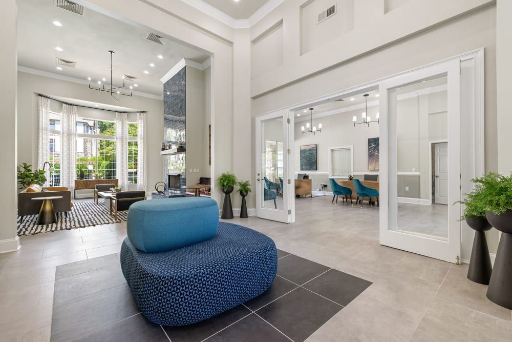 Spacious modern lobby area with high ceilings, tiled floors, and large windows allowing abundant natural light. The room features stylish furniture including a large blue ottoman, chic armchairs, and a black and gold side table. Two sets of French doors open into adjacent rooms, with one revealing a dining area with a long table and teal chairs at Marquis Carmel Commons in Charlotte, NC.