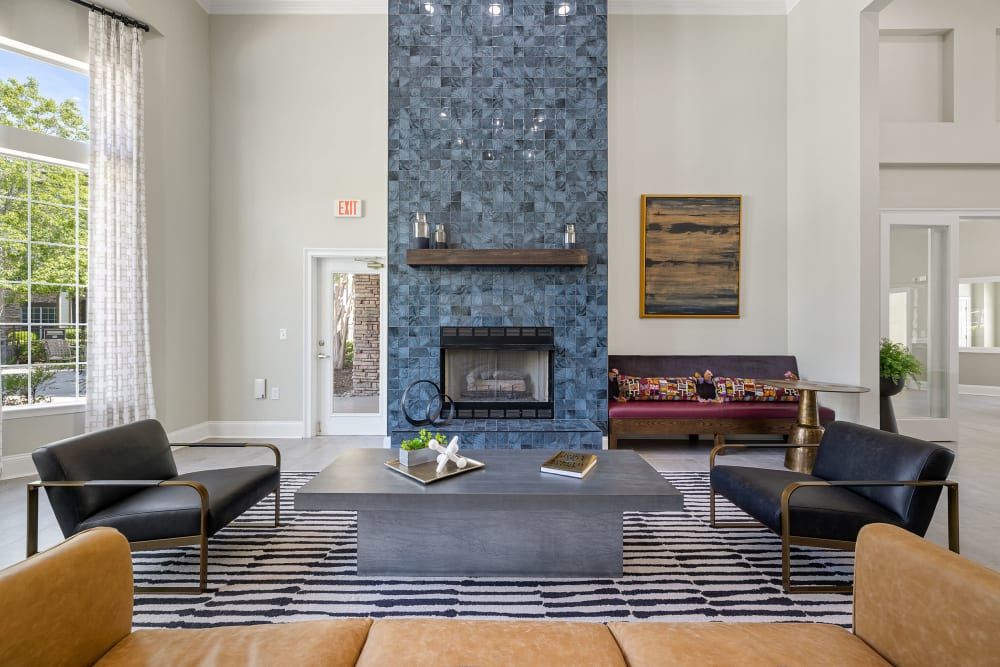 Spacious living room with high ceilings, a blue tiled fireplace, modern furniture, including a tan sofa, black chairs, and a dark coffee table on a patterned area rug. A landscape painting hangs above a bench on the right, with natural light coming from windows with sheer curtains at Marquis Carmel Commons in Charlotte, NC.