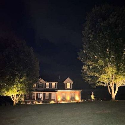 A house at night, lit by spotlights. Trees flank either side on a grassy lawn under a dark sky.