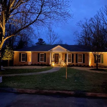Lit-up brick house at dusk, with lights highlighting the facade and trees on a green lawn.