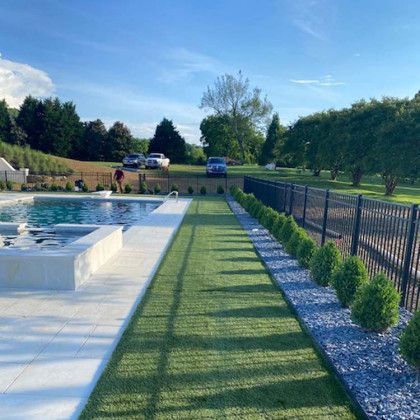 Poolside view with pool, artificial turf, black fence, trimmed bushes, and cars in the distance.