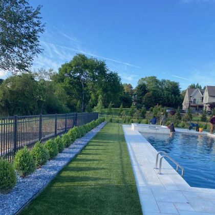 Poolside view with grass, shrubs, and a fence. Blue sky, water, and trees in the background.