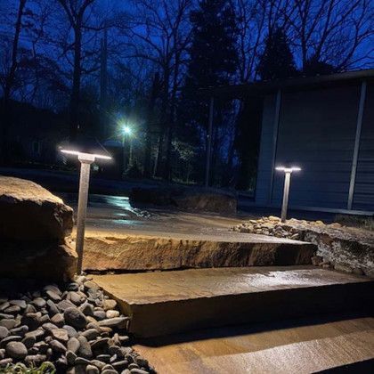 Outdoor pathway lit by two modern lights, stone steps, dark trees in the background, night scene.