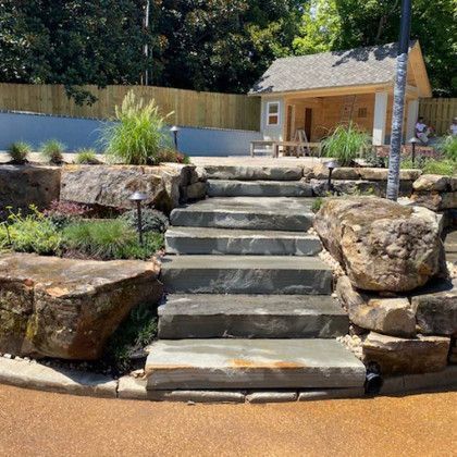 Stone steps lead to a small wooden building, surrounded by landscaping and retaining walls.