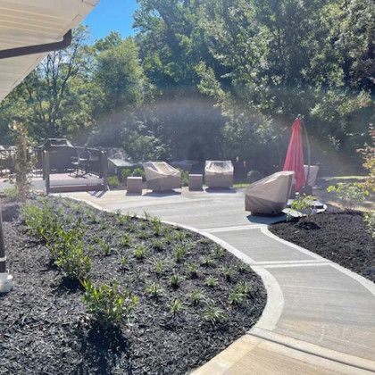 Concrete patio with seating area, pathway, and landscaping with mulch and bushes, sunny outdoor setting.