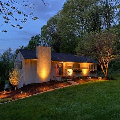 Cottage illuminated at dusk with warm lights, chimney, flag, flower boxes, and a grassy hill in the background.
