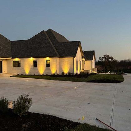 Large white house with dark roof, lit up with warm exterior lighting, on a concrete driveway.