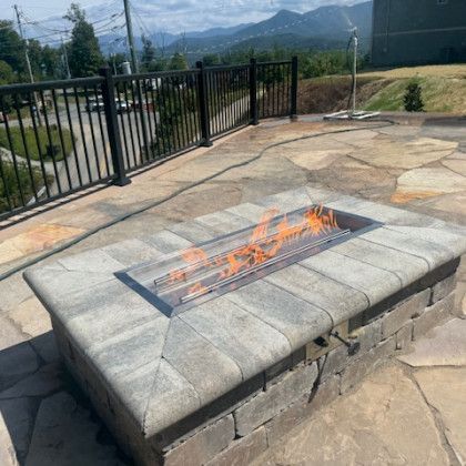 Fire pit with flames on a stone patio, black railing, mountain view in background.