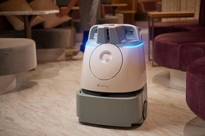 Small white robot with a blue light, sitting on a floor in a modern lounge.