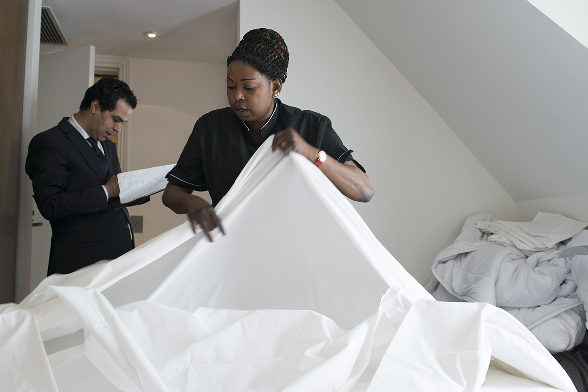 Two hotel staff making a bed with white sheets in a bright room