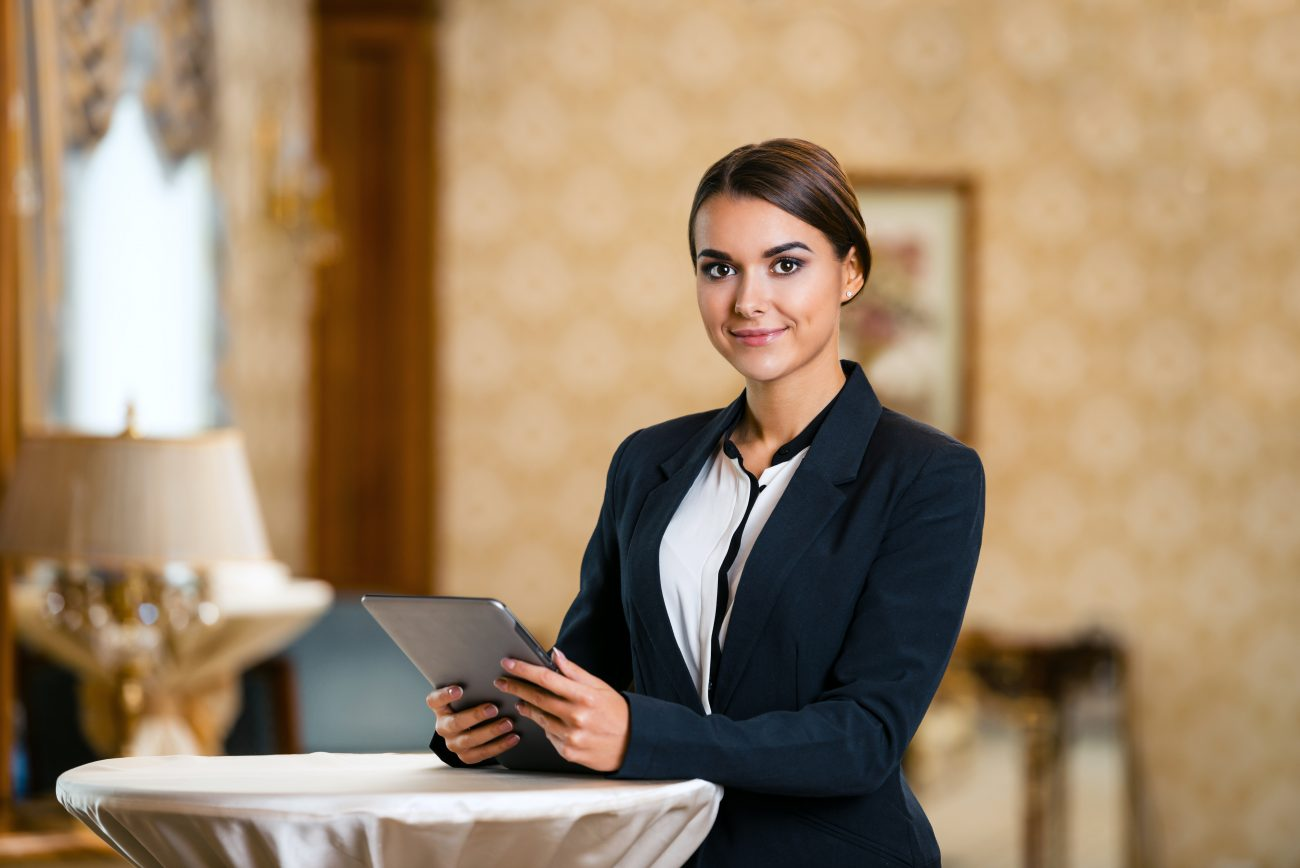 Smiling hotel receptionist in a dark blazer holding a tablet behind a table in a softly lit lobby