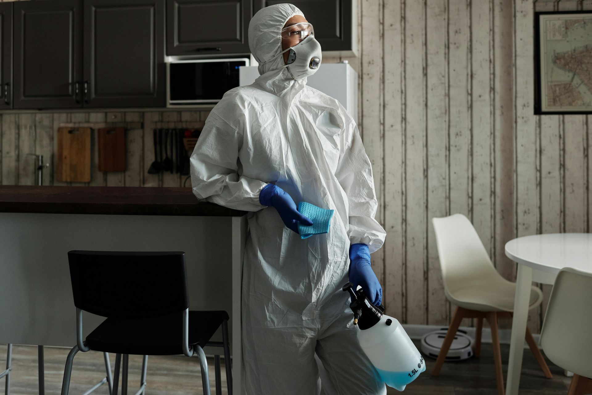 Person in white protective gear and blue gloves holding a sprayer in a kitchen.