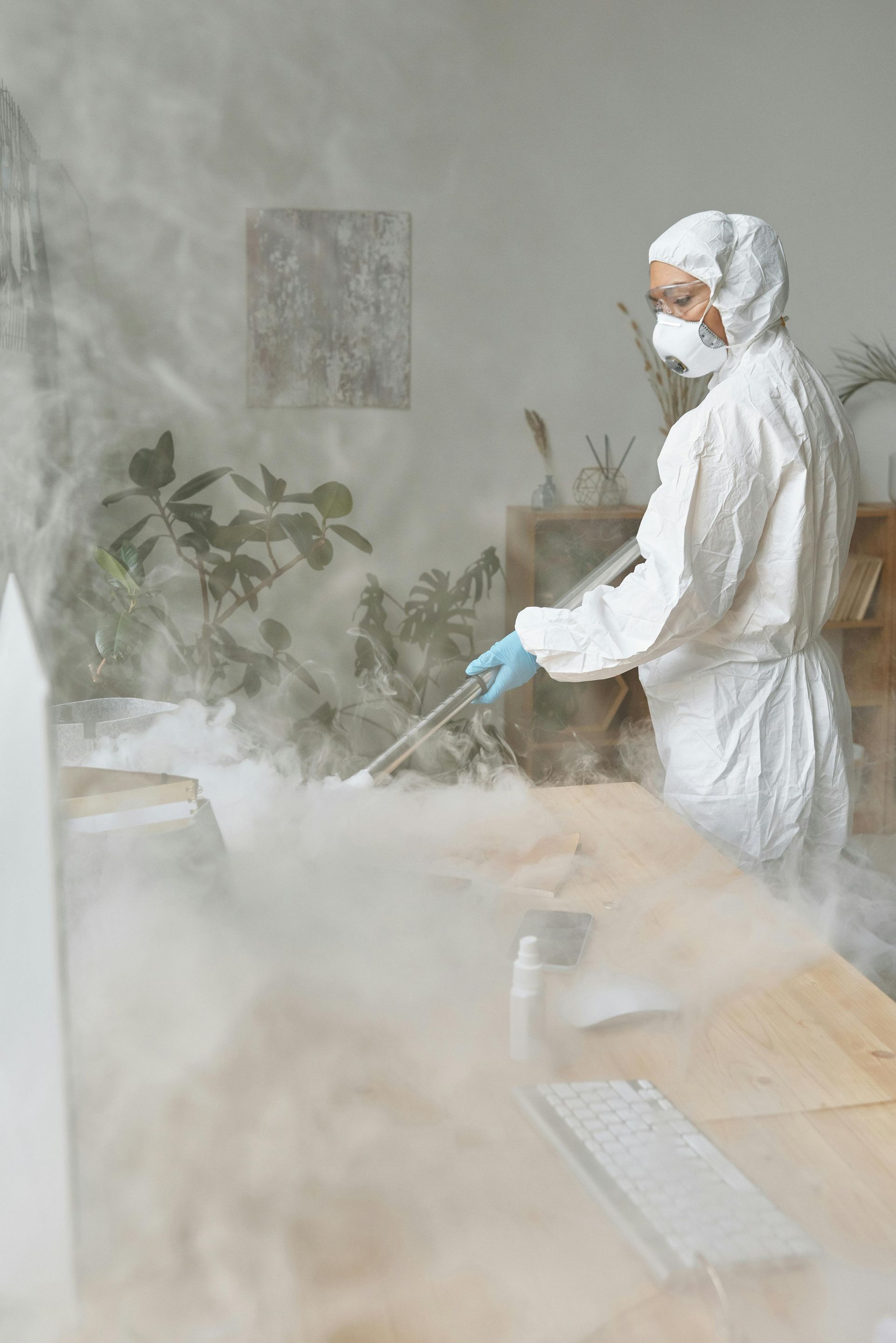 Person in protective suit spraying disinfectant in a hazy room with plants and a computer monitor