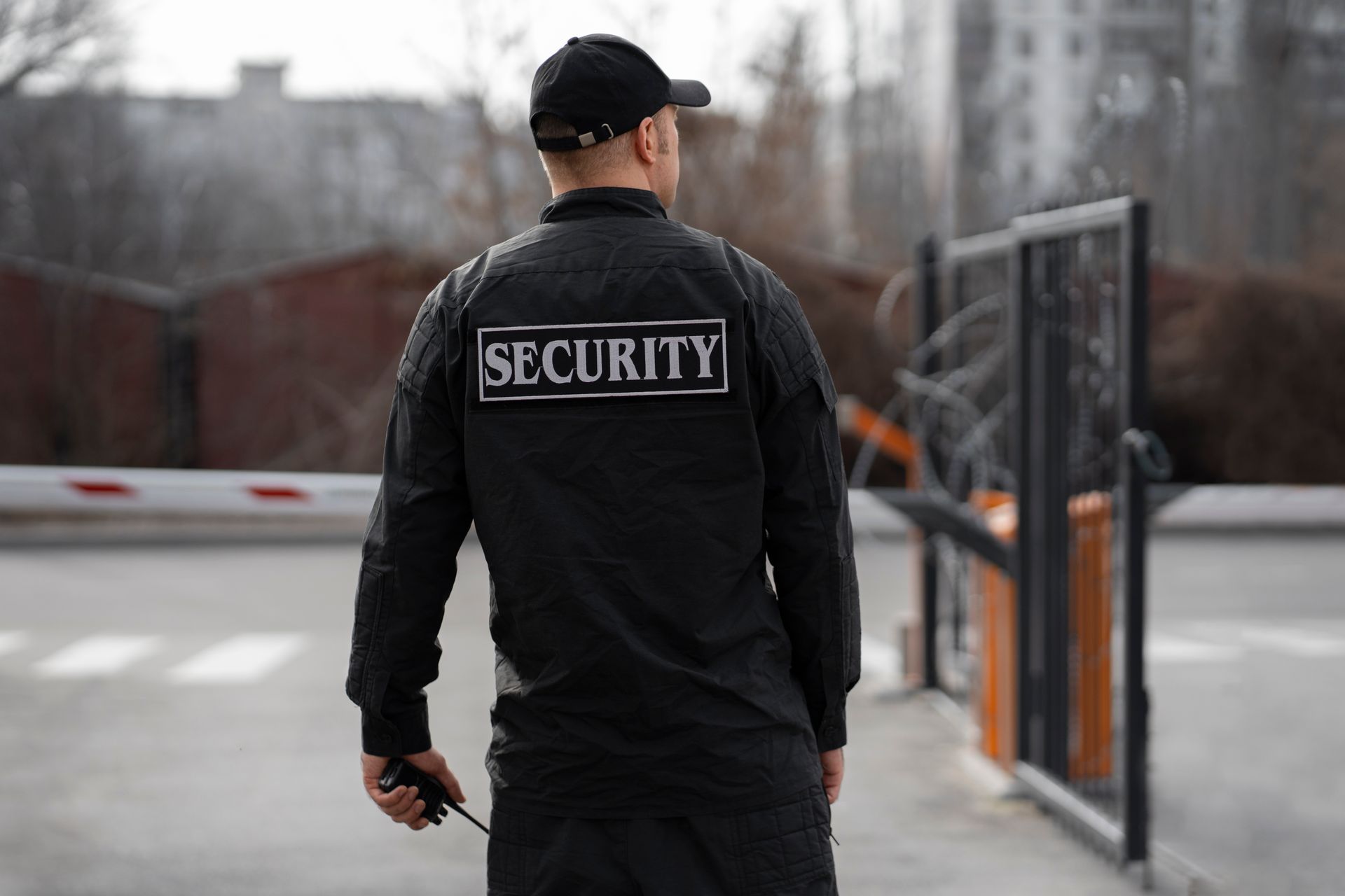 Security guard standing at an entrance gate, facing away in black uniform with “SECURITY” on back.