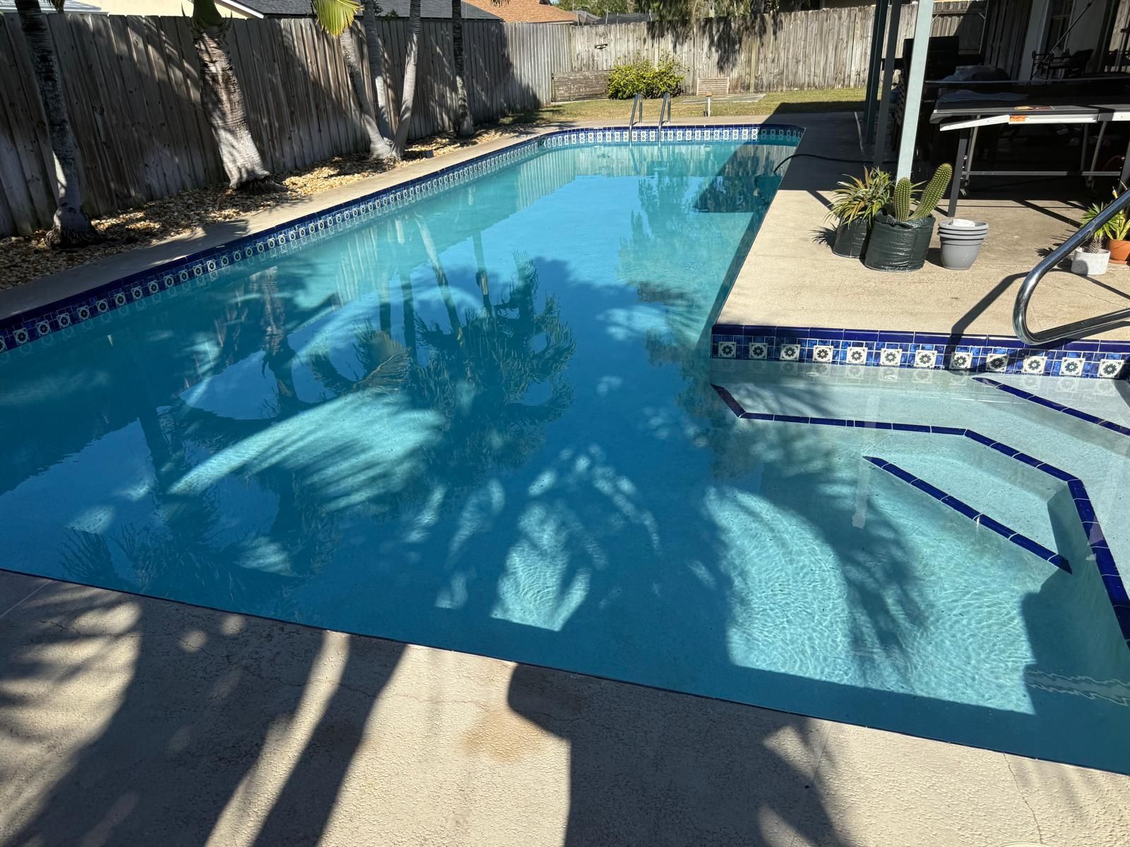 A rectangular swimming pool filled with clear water, with steps at one end. Palm tree shadows reflected.