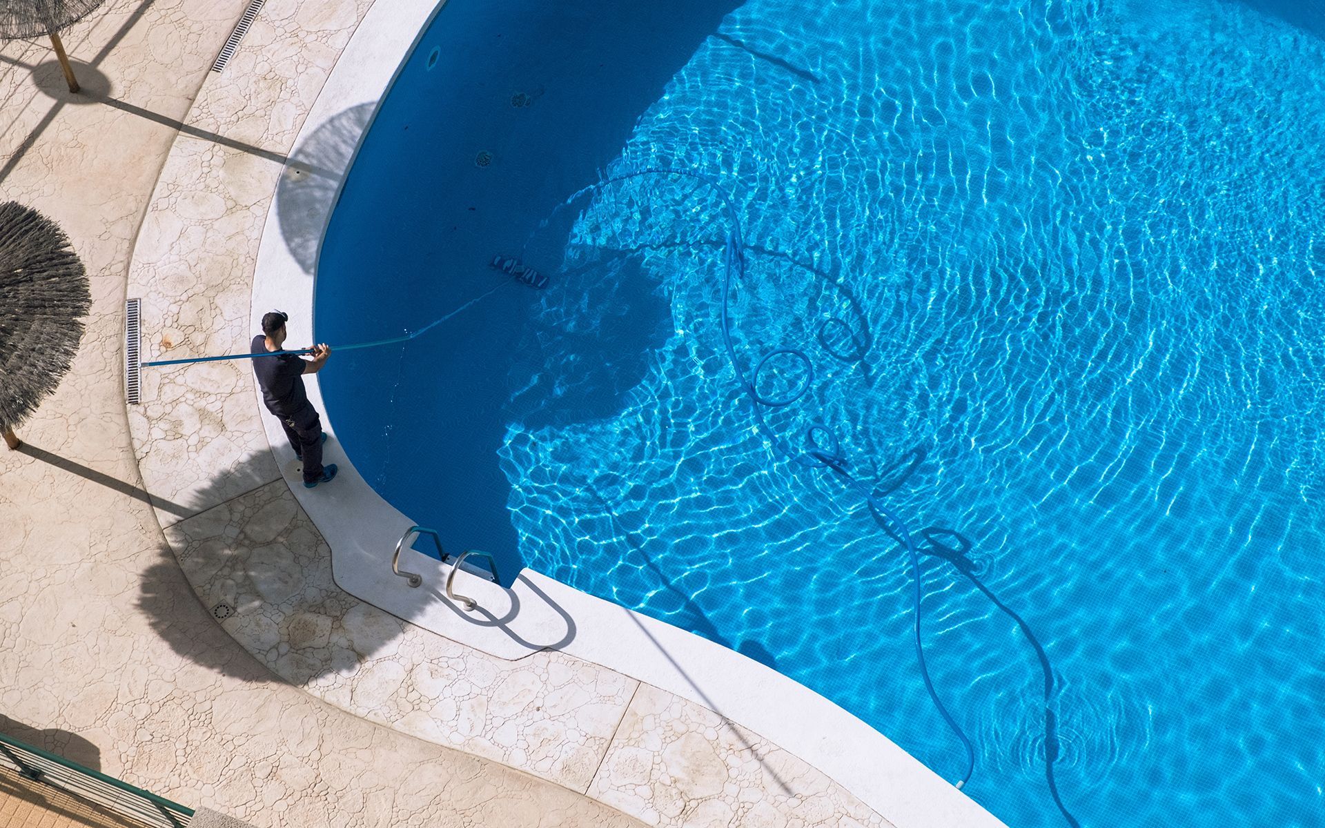 Person cleaning a blue, kidney-shaped swimming pool with a long brush on a sunny day.