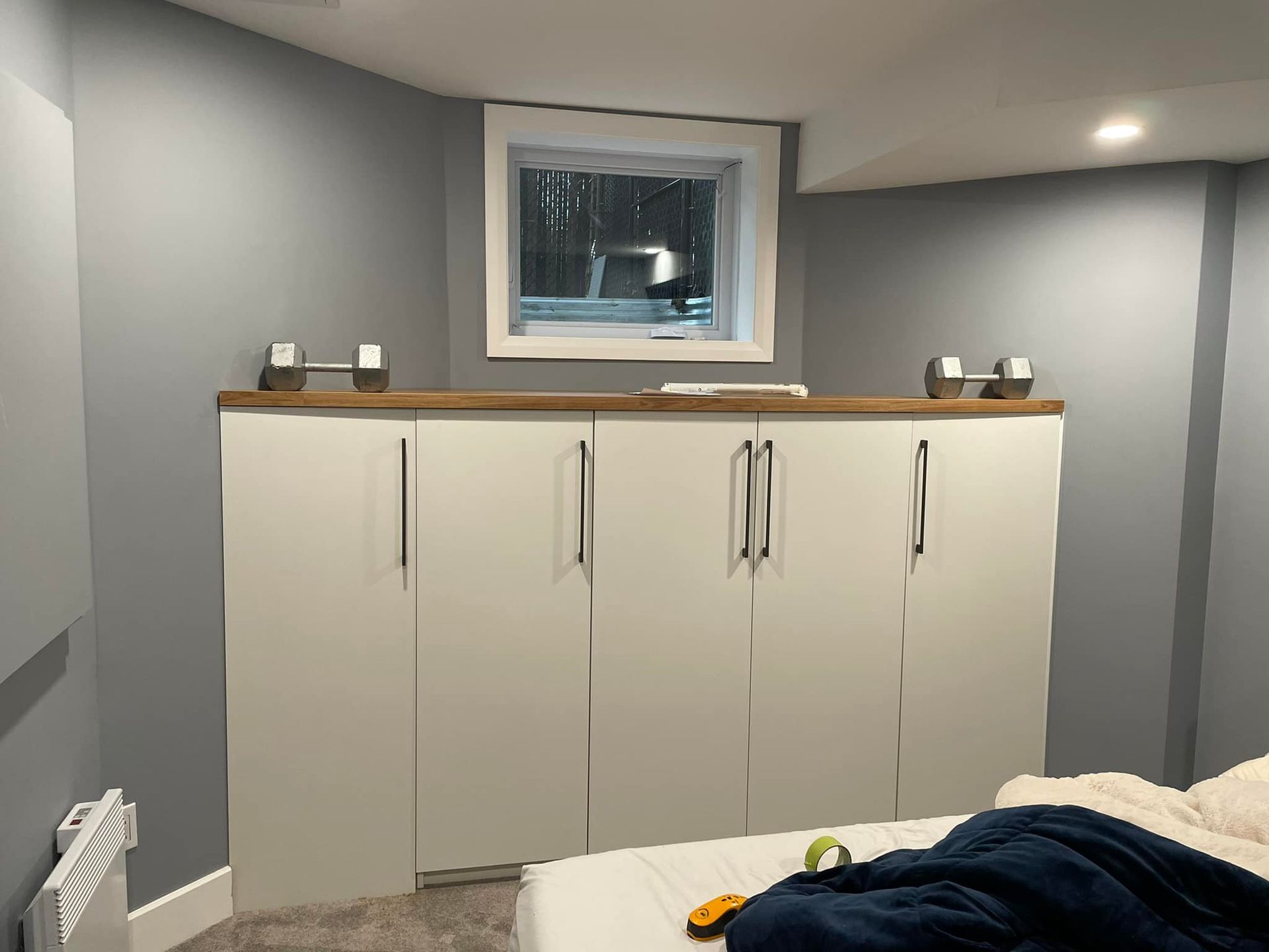 Chambre grise avec des armoires blanches sous une petite fenêtre et un bord de lit au premier plan