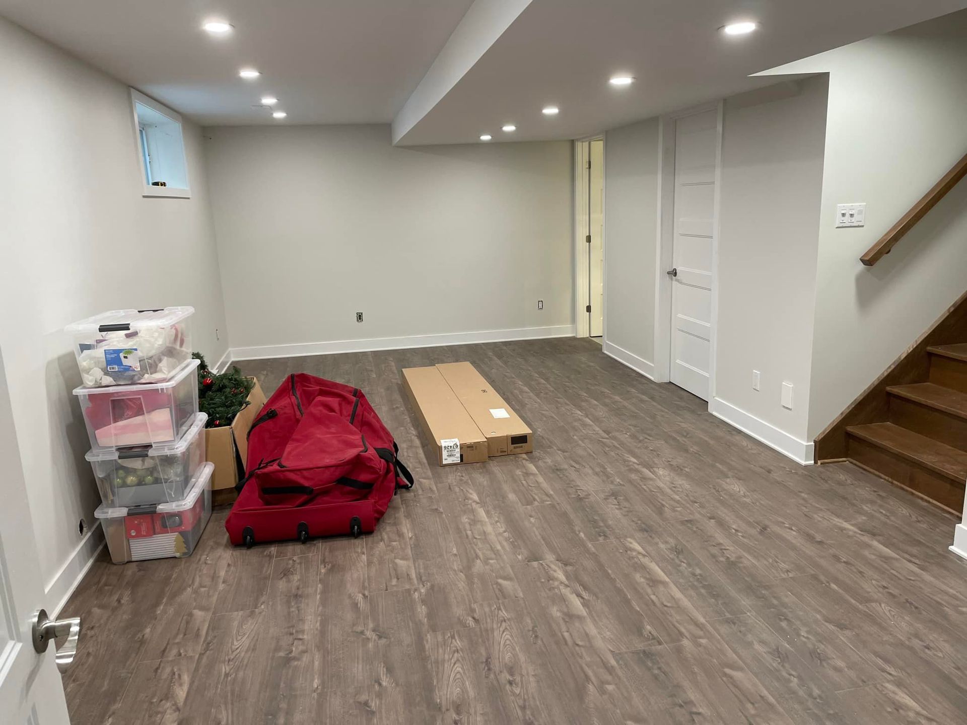 Sous-sol non aménagé avec plancher en bois, bacs de rangement empilés, pouf rouge, boîte en carton et escalier sur la droite.