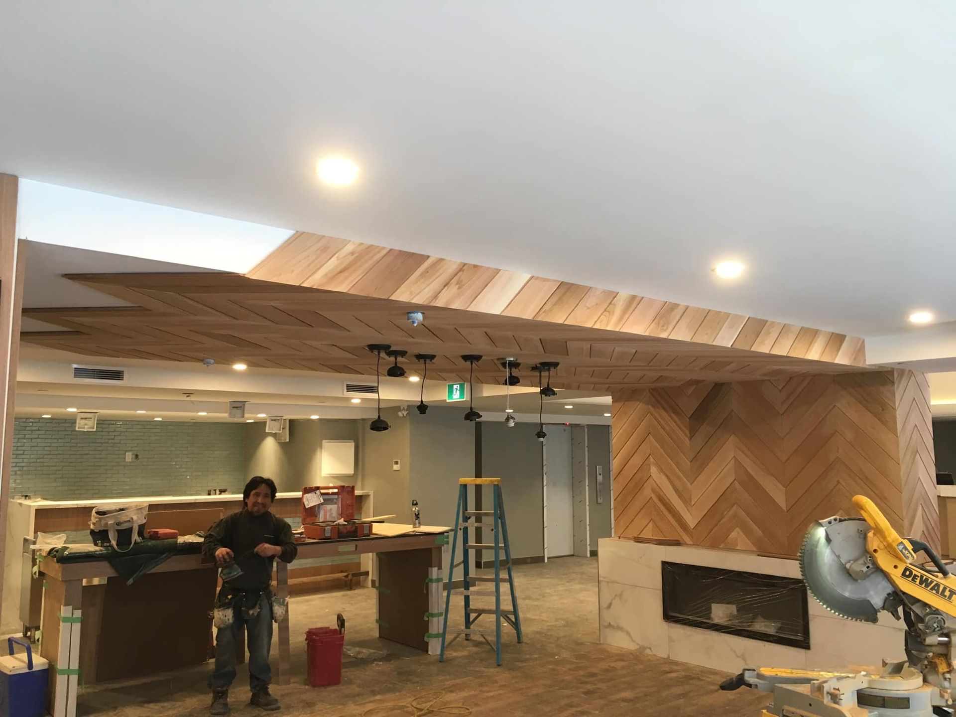 Des ouvriers du bâtiment installent un plafond en bois à chevrons dans une pièce moderne à l'aide d'outils et d'une échelle.