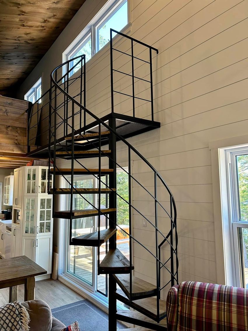 Un escalier intérieur en colimaçon mène à une mezzanine dans une pièce lumineuse aux murs en bois et aux grandes fenêtres.
