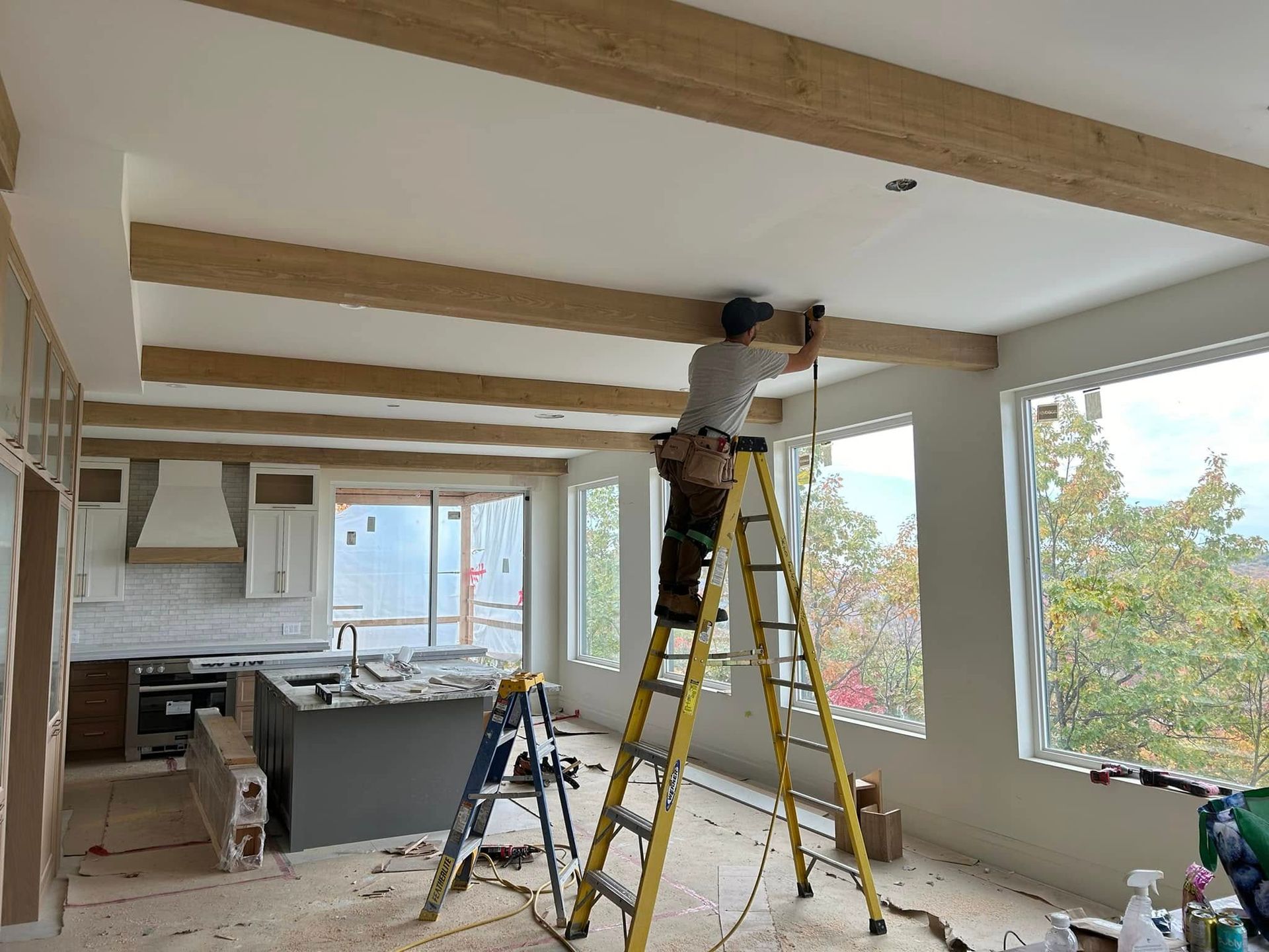 Une personne sur une échelle installe des luminaires au plafond dans une cuisine lumineuse et inachevée avec des poutres apparentes.