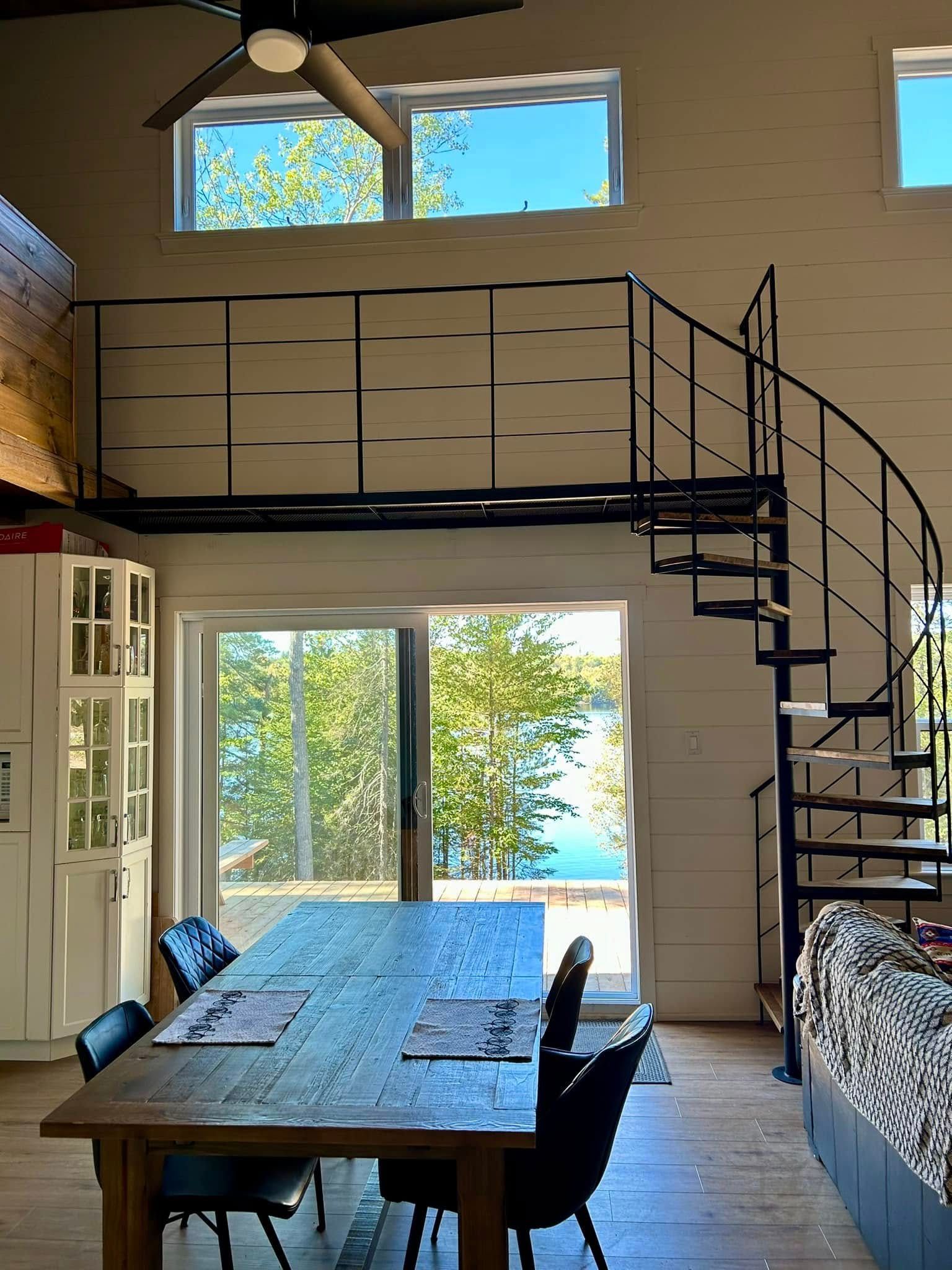 Coin repas en mezzanine avec table en bois, escalier en colimaçon, hautes fenêtres et vue sur les arbres et l'eau