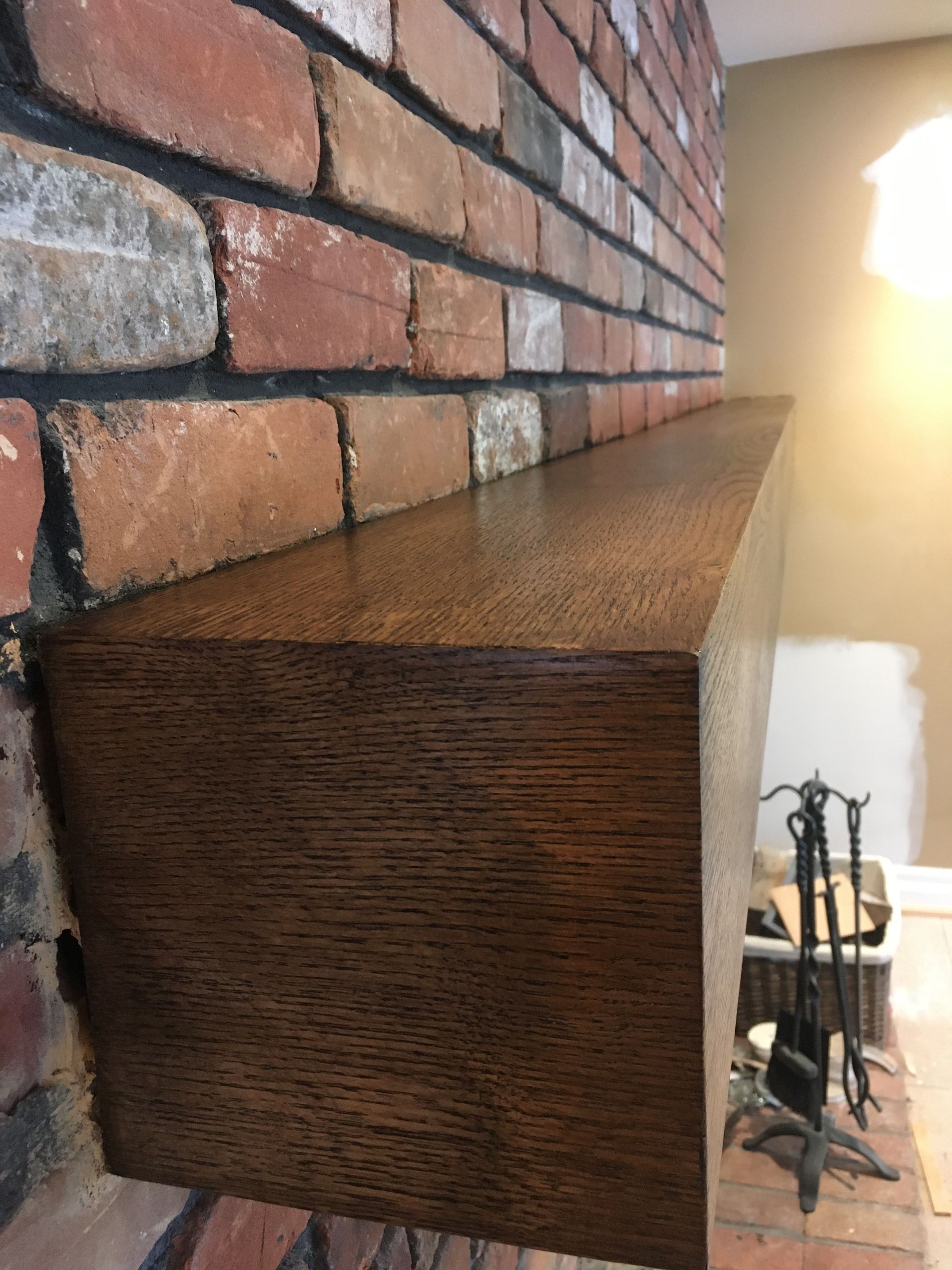 Étagère de cheminée en bois brun contre un mur de briques, baignée de lumière naturelle, avec un ensemble d'accessoires de cheminée en dessous.