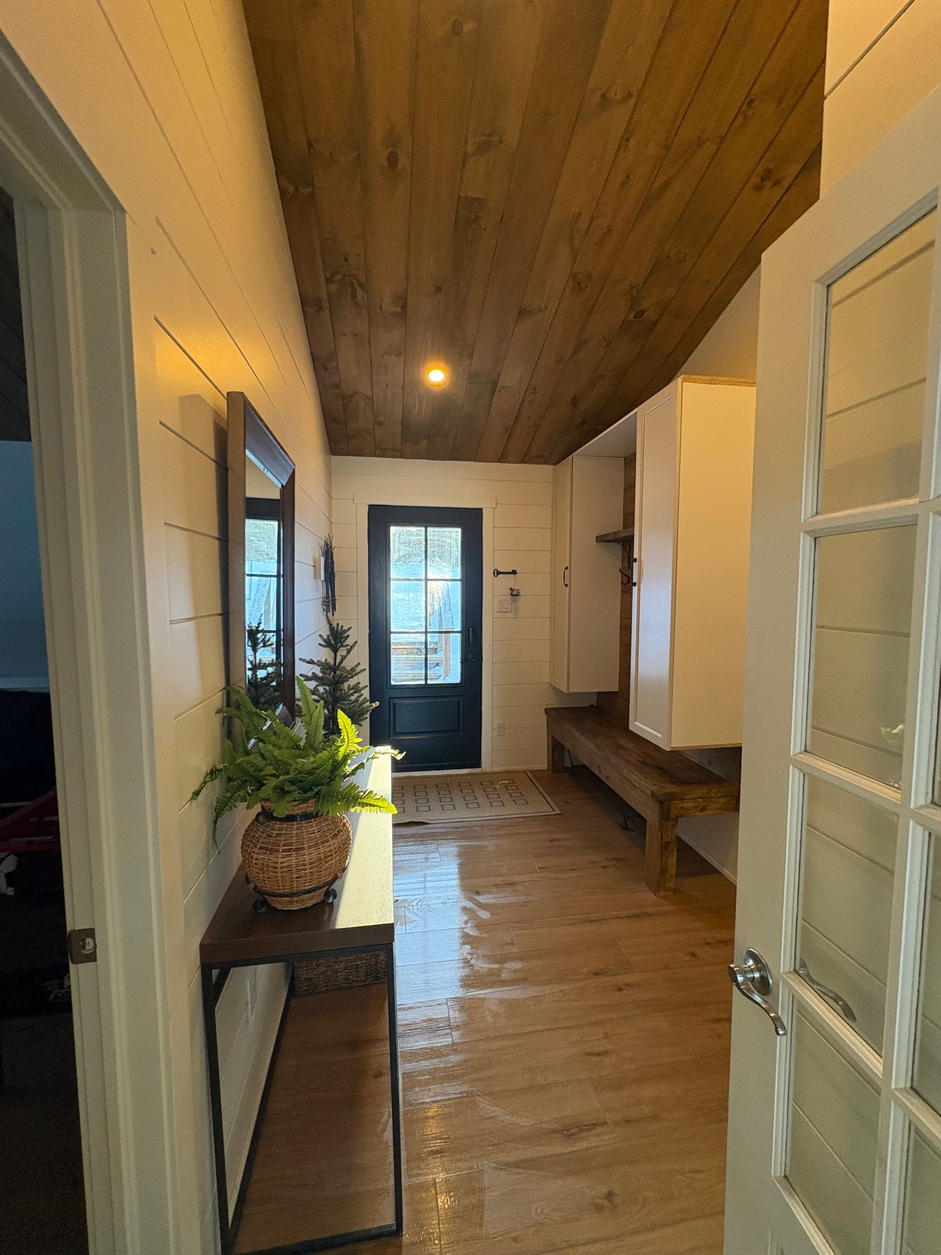 Couloir étroit avec plafond et sol en bois, murs blancs, banc, plante et porte vitrée au fond.