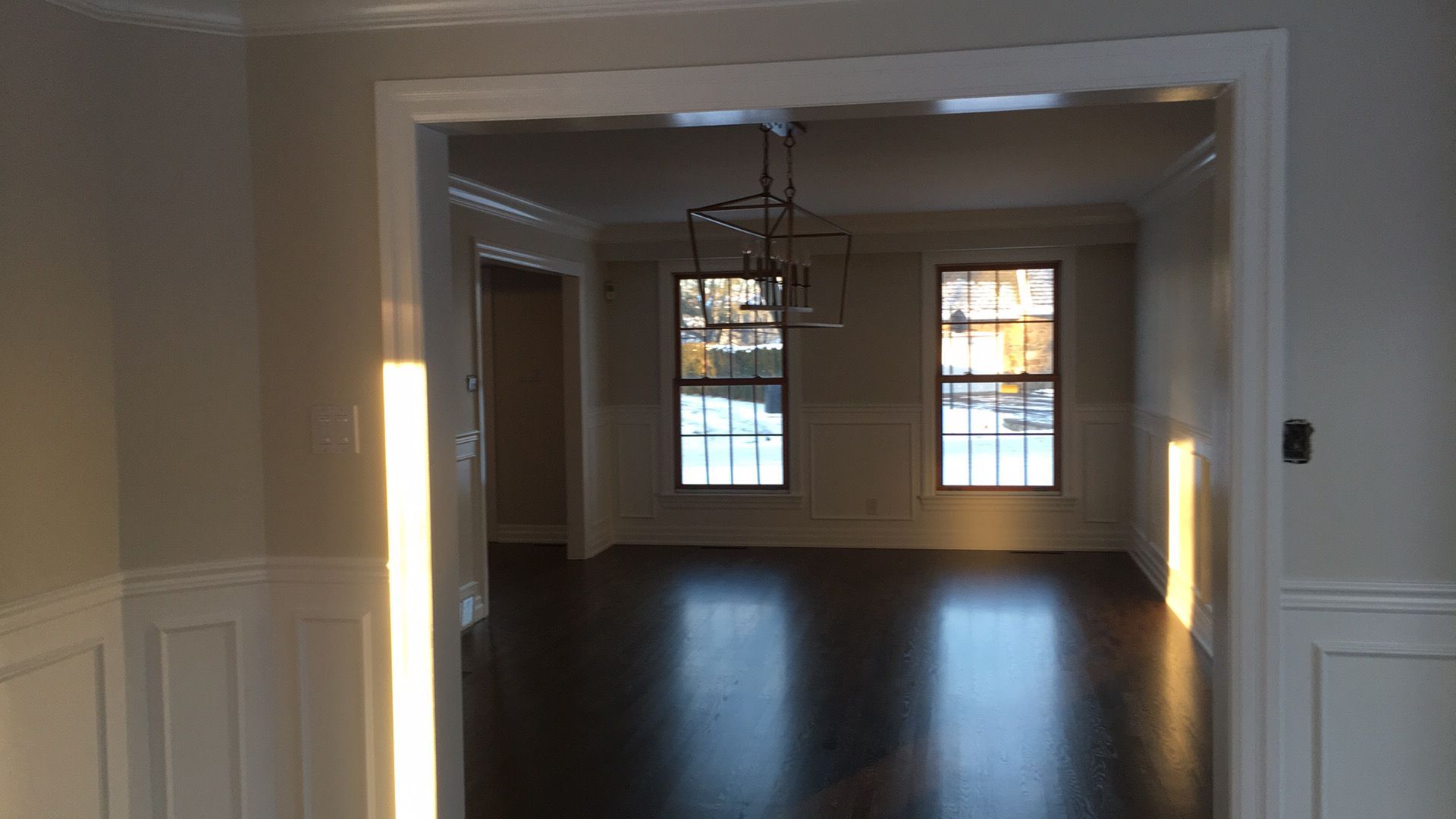 Salle à manger vide avec parquet, murs blancs et lustre, visible à travers une large porte.