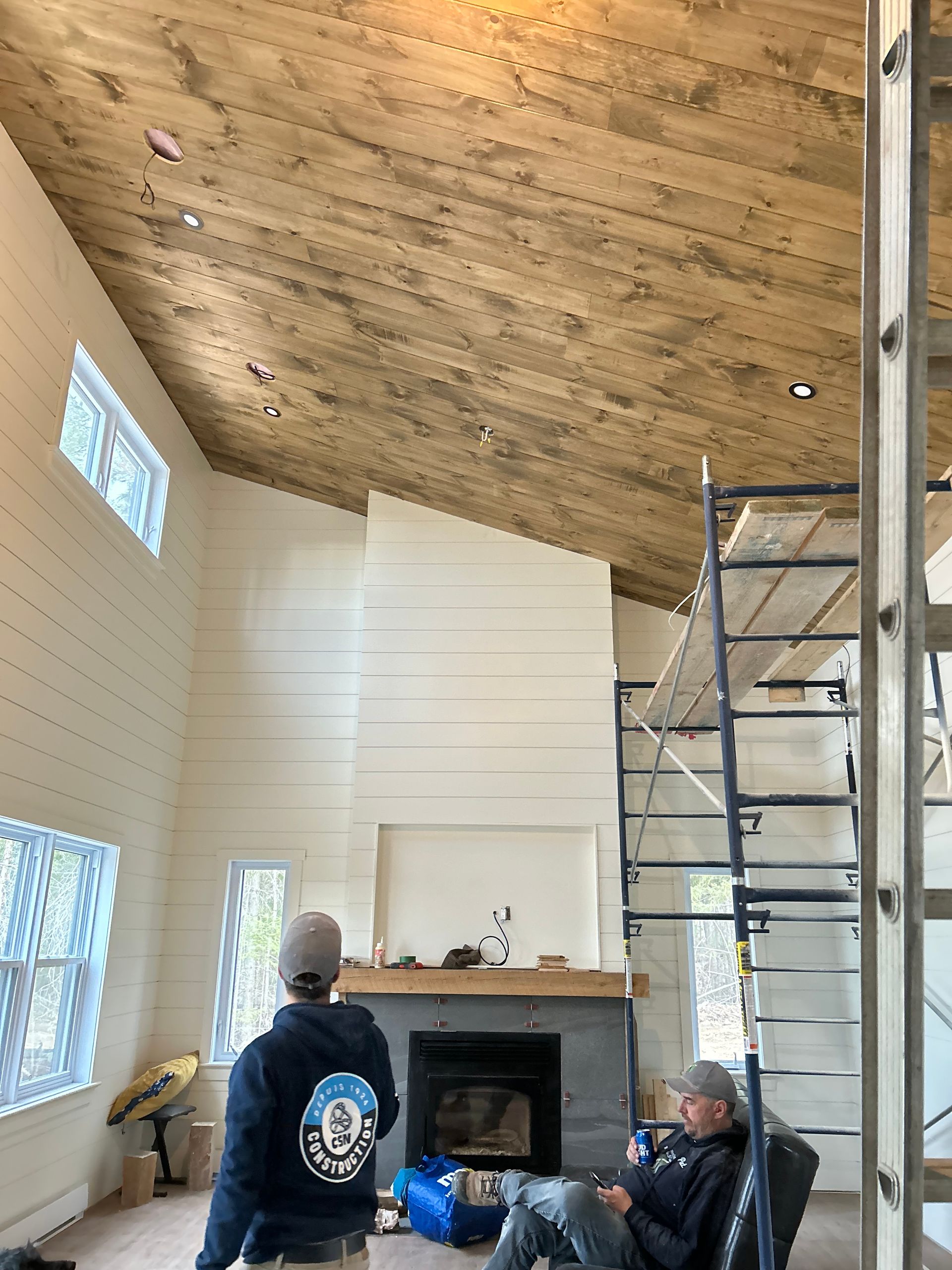Deux personnes dans une cabane en construction, avec une cheminée en pierre, un plafond en bois et des échafaudages.