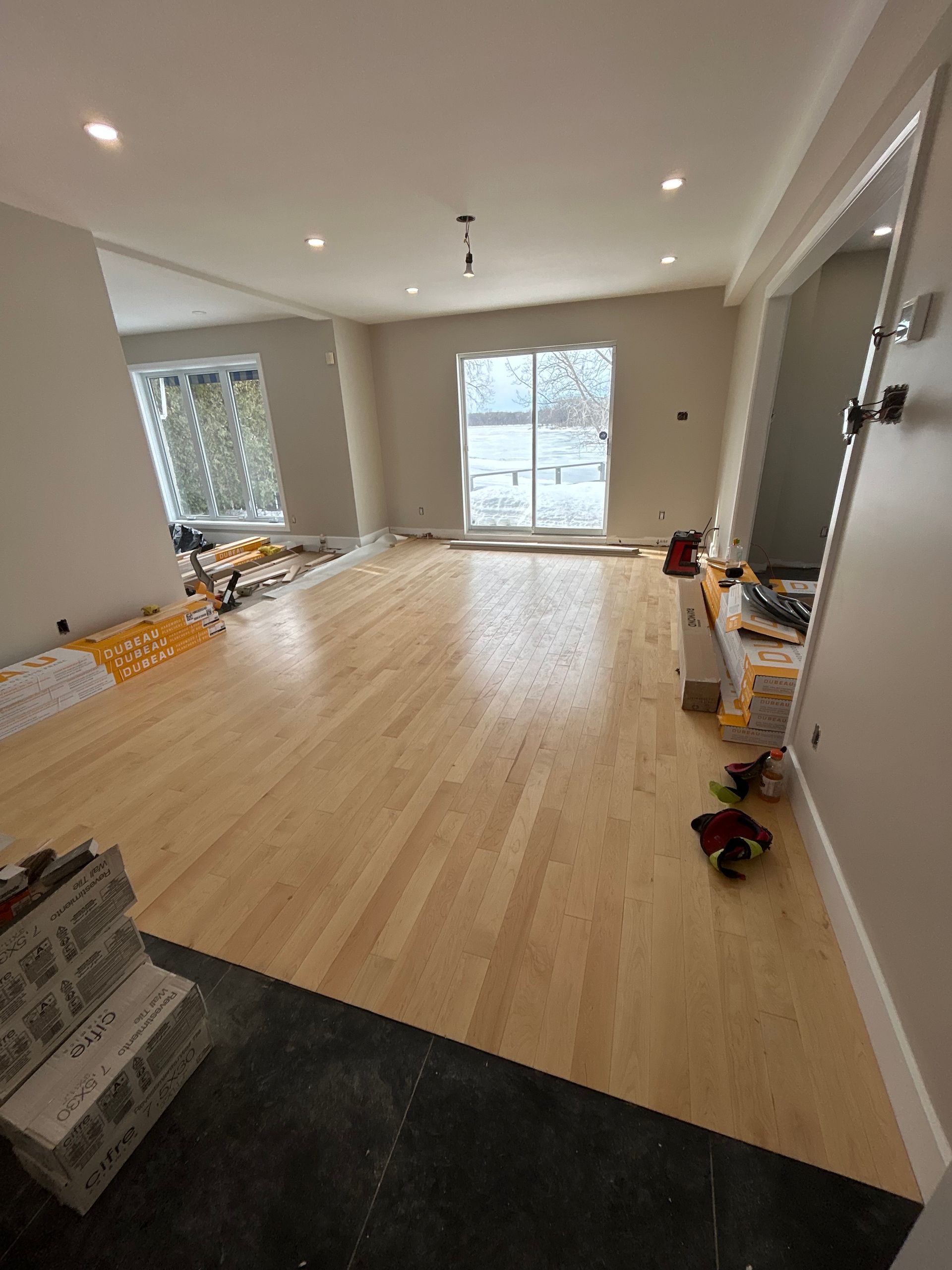 Salon vide avec parquet clair, spots encastrés et grandes fenêtres.