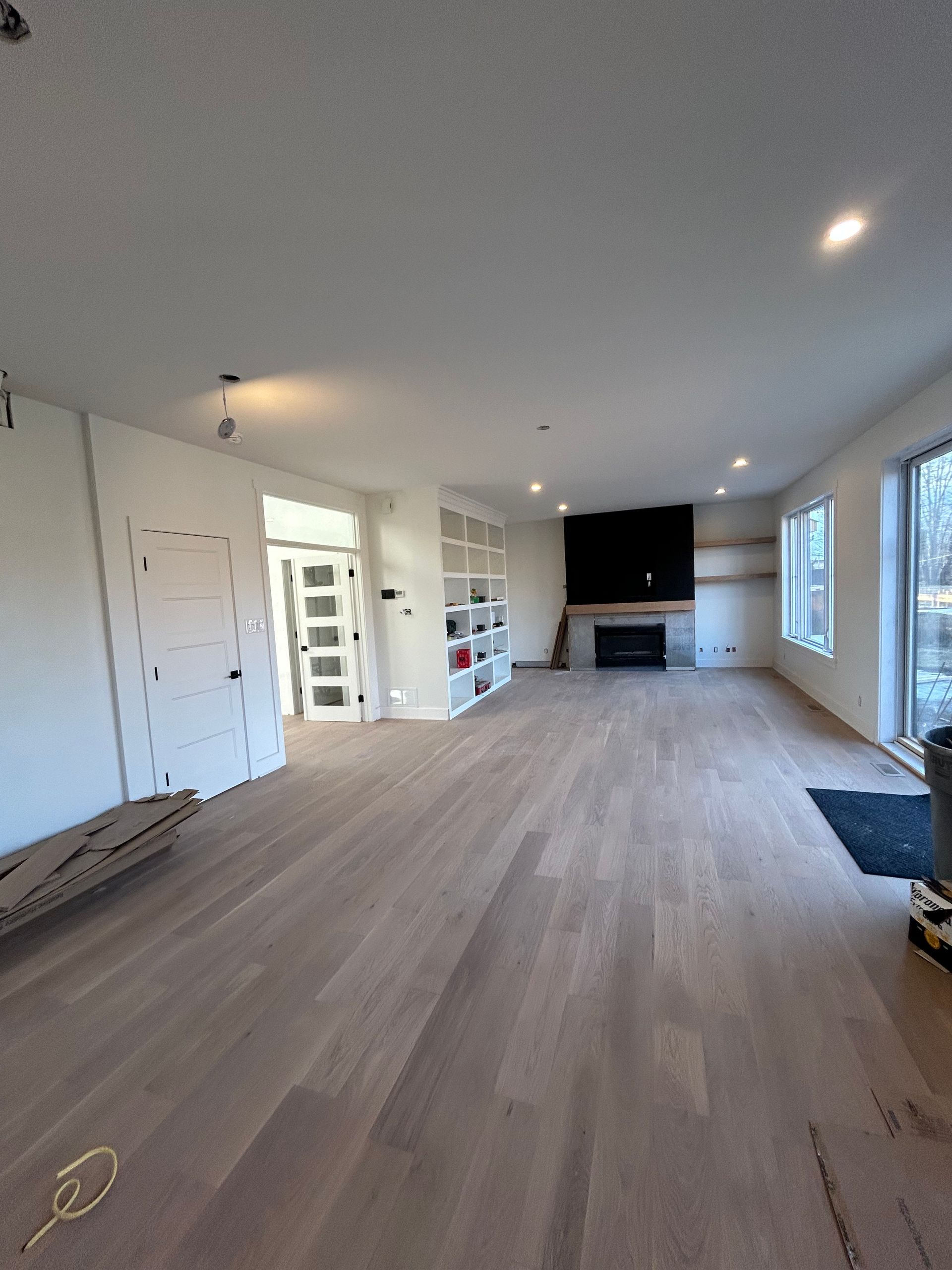 Salon moderne vide avec parquet clair, murs blancs, cheminée et grandes fenêtres.