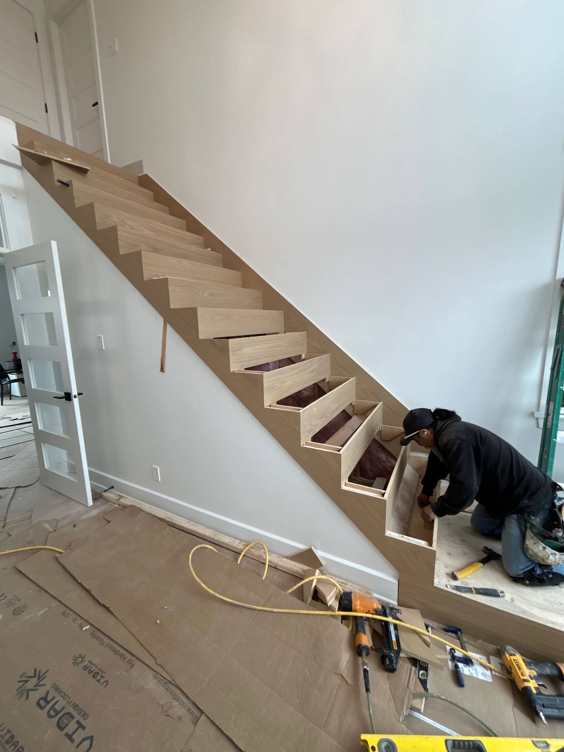 Un ouvrier installe un escalier flottant en bois dans une pièce inachevée, à l'aide d'outils et de matériaux de construction.