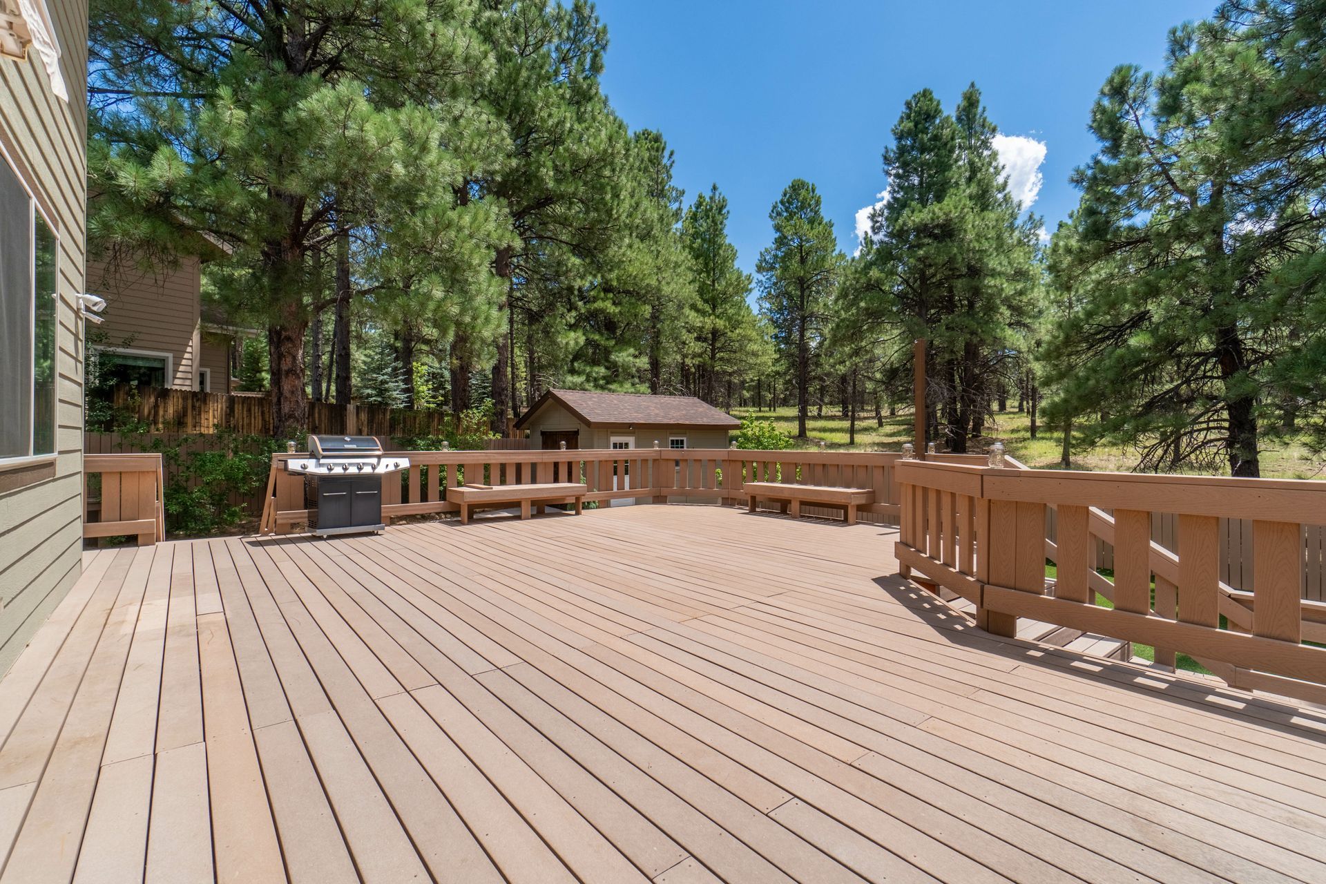 Spacious wooden deck with railings, hot tub, and pine trees under a clear blue sky