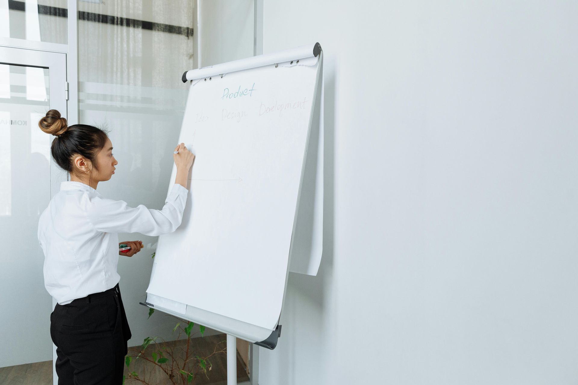 A person in a white shirt and black pants stands at a flip chart, writing on the paper in a brightly lit office.
