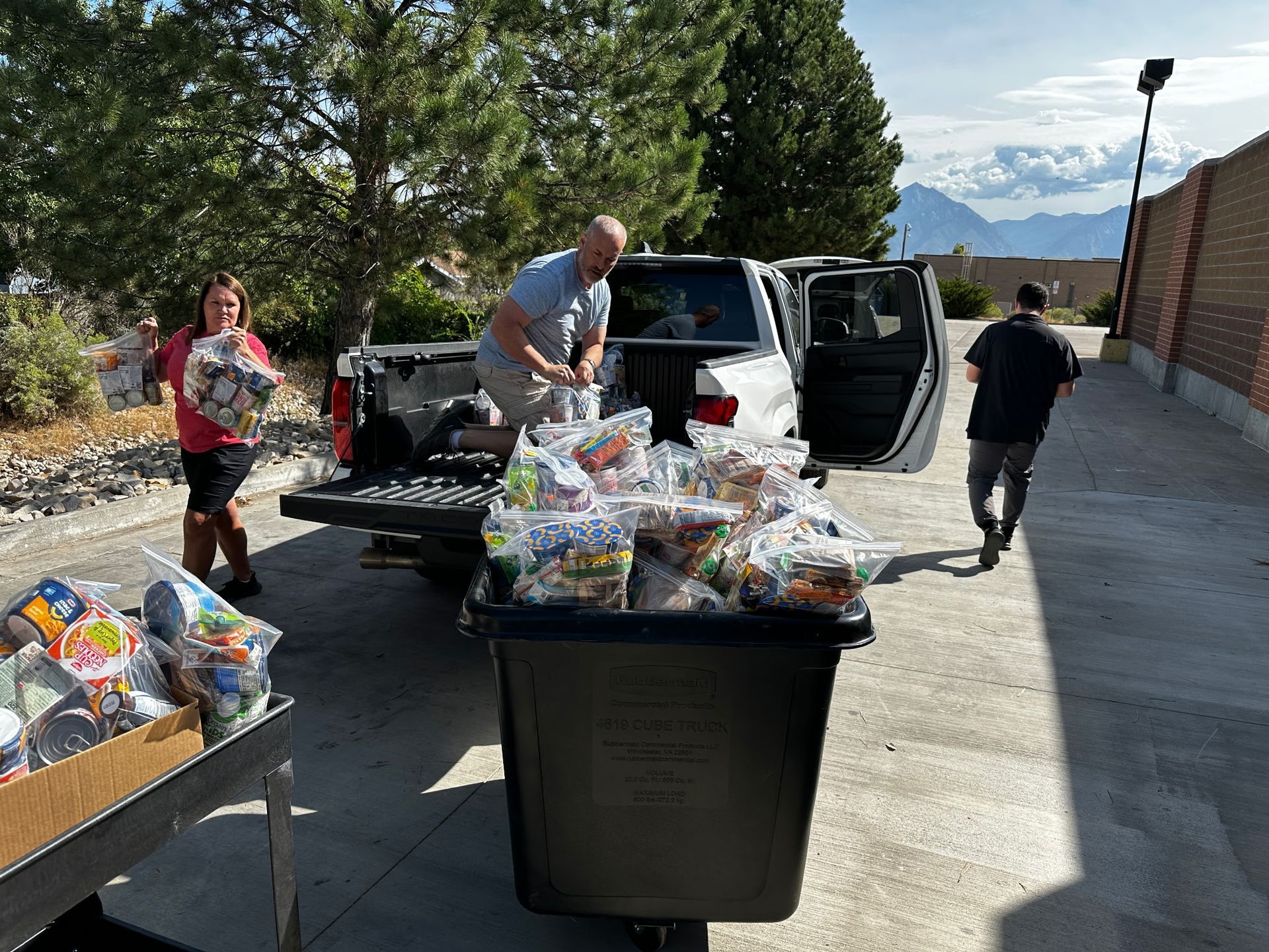 People unloading bags of food from a truck into a large container outdoors.
