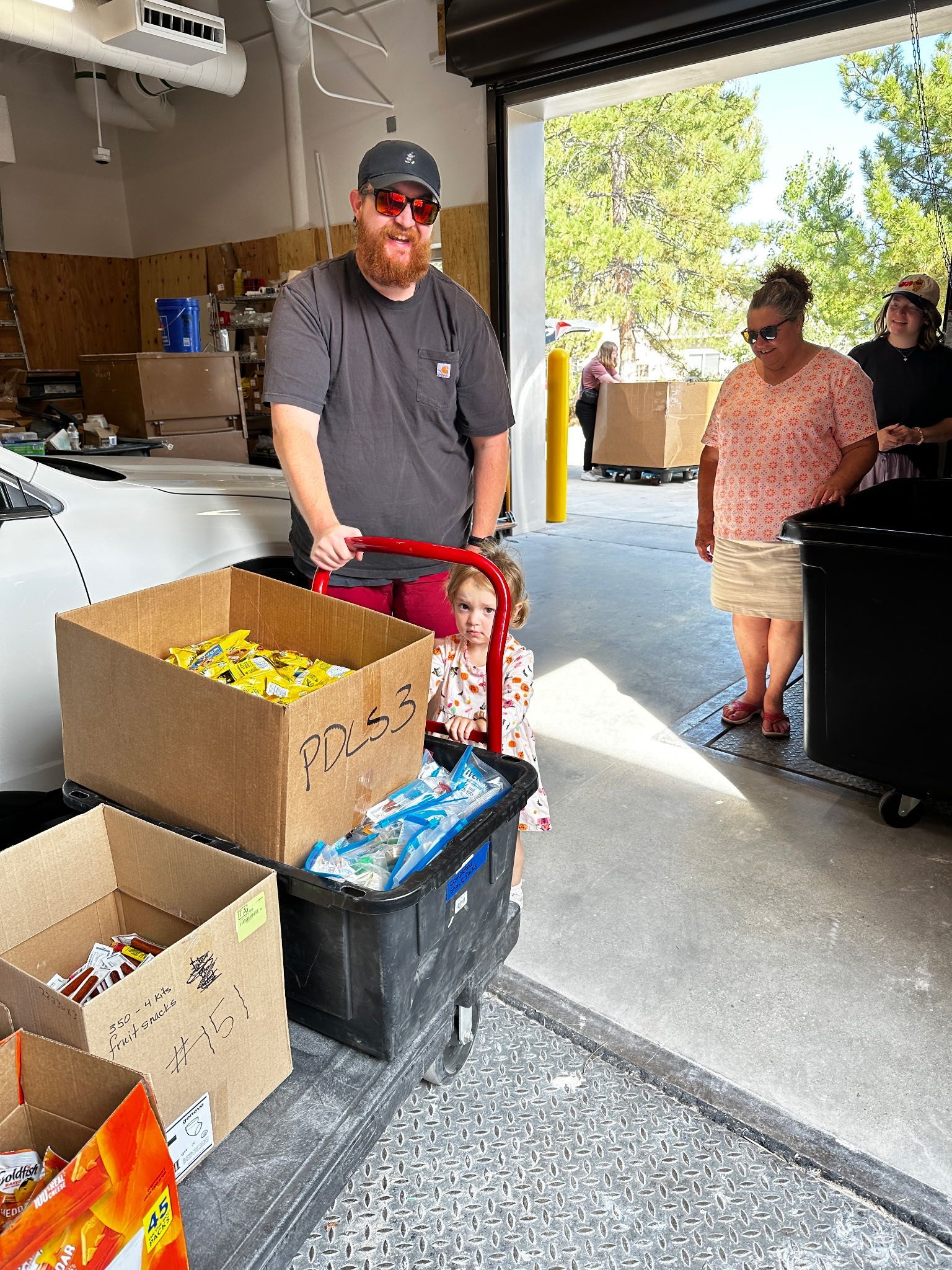 Man and child load supplies on a cart, inside a warehouse. Other people stand nearby.