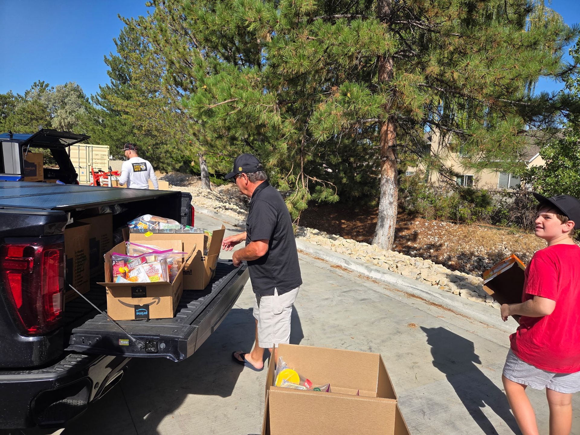 People unloading boxes from a pickup truck, likely for a food drive, sunny outdoor setting.