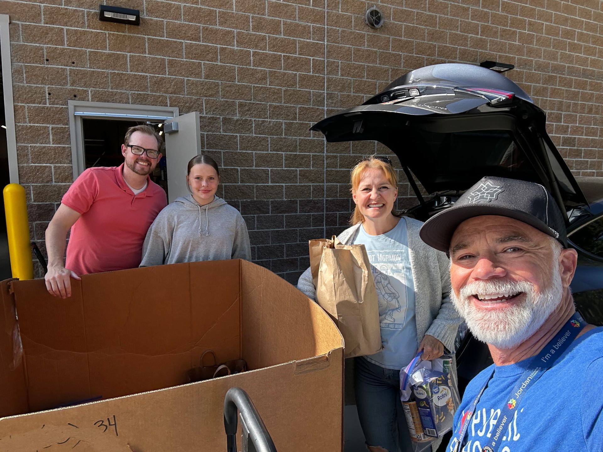 Four people loading a car with food donations, smiling. Outdoors, brick building.