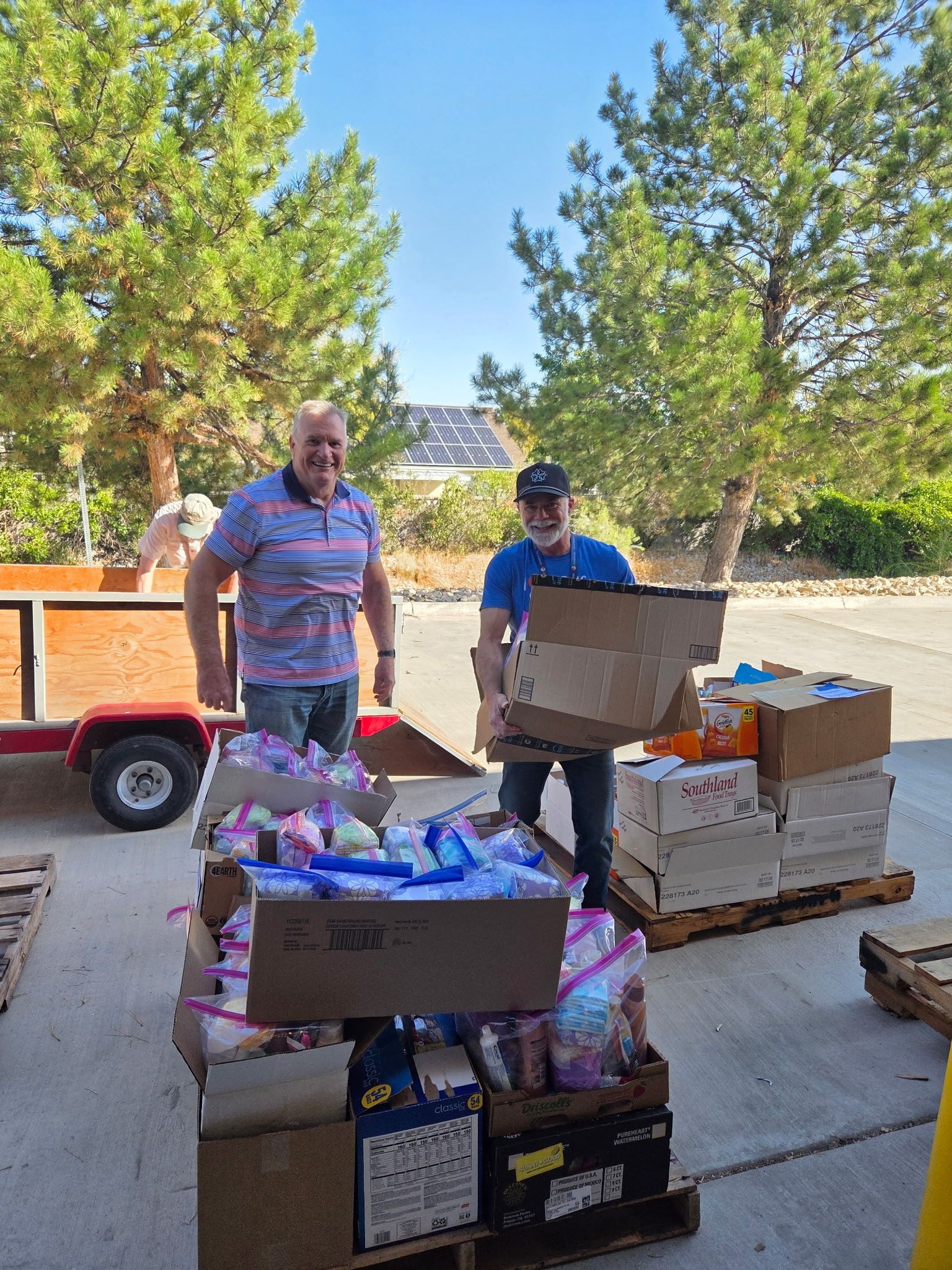 Two men loading boxes of donations onto pallets outside on a sunny day.