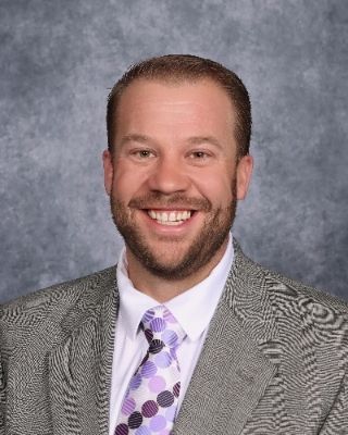 Man wearing a suit and tie, smiling at the camera against a neutral background.