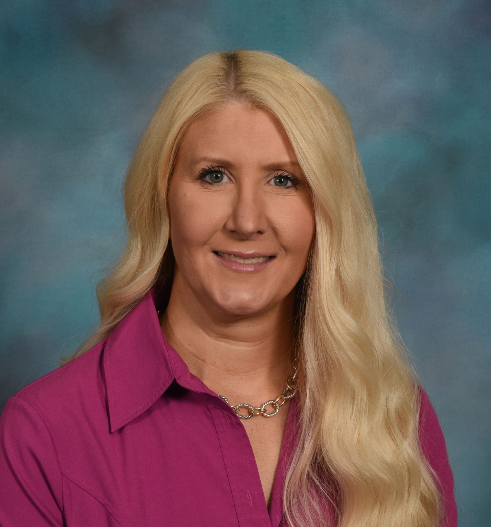 A woman with long blonde hair is wearing a pink shirt and a necklace.